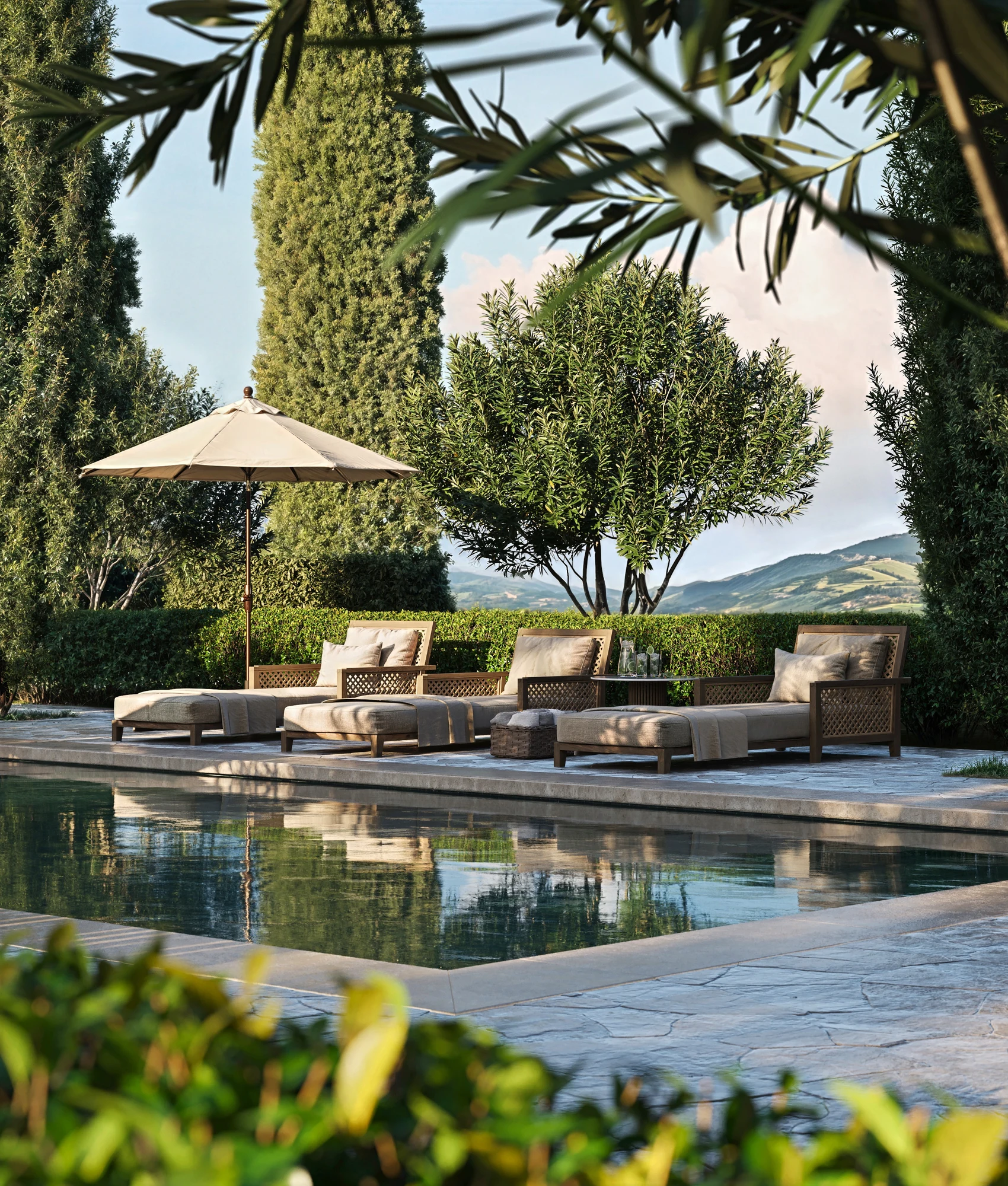 Poolside loungers beneath a parasol, framed by foliage and cypress trees